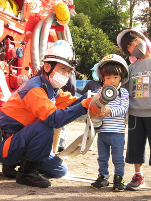 ヘルメットを付けて消防士の方と一緒にホースを持っている男の子の写真