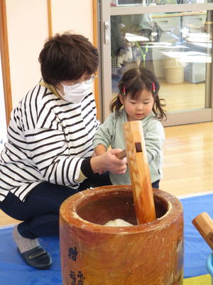 女性と一緒に杵を持ち臼の中の餅をついている女の子の写真