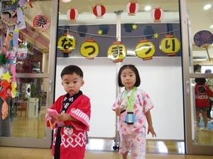 法被を着た男の子と甚平を着た女の子がなつまつりの飾り付けの前に立っている写真