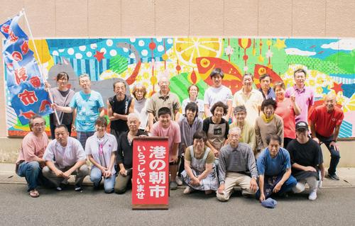 港の朝市いらっしゃいませと書かれた看板を中央に、市場関係者が20名ほど3列で並んでいる写真