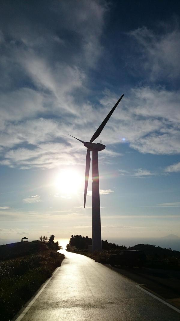 うす暗い中、奥まで続く一本道沿いに立つ風力発電所の合間から日の光がみえ、うつろう空が綺麗なグラデーションに染まっている写真