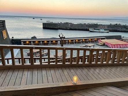オレンジがかった空、海に浮かぶ堤防に人影が見えており、手前にはムーンロードテラスに繋がる木材でできたスロープが写っている写真
