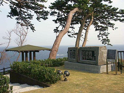 海を背景に、幹の太い木が3本生えている芝生の公園に、大きな石の歌碑がある写真